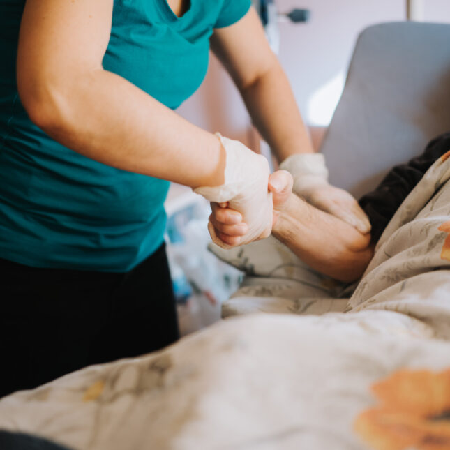 A person, wearing gloves, holds the hand and arm of a patient who lays on a hospital bed — in the lab coverage from STAT