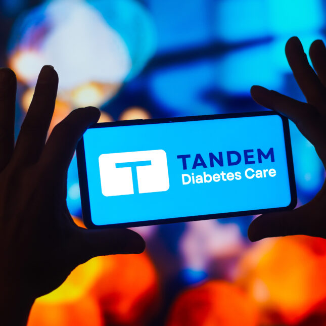 Two hands hold a phone, of which the screen displays Tandem Diabetes Care logo, in front of a blue and orange projection — coverage from STAT