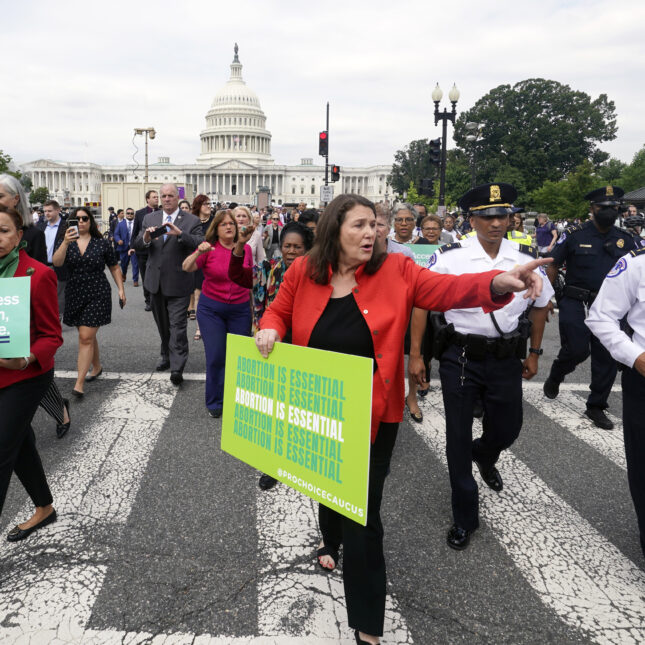 Rep. Diana DeGette holds a sign that repeats "ABORTION IS ESSENTIAL" five times, pointing to her left on a crosswalk, while marching from the Capitol to the Supreme Court — first opinion coverage from STAT