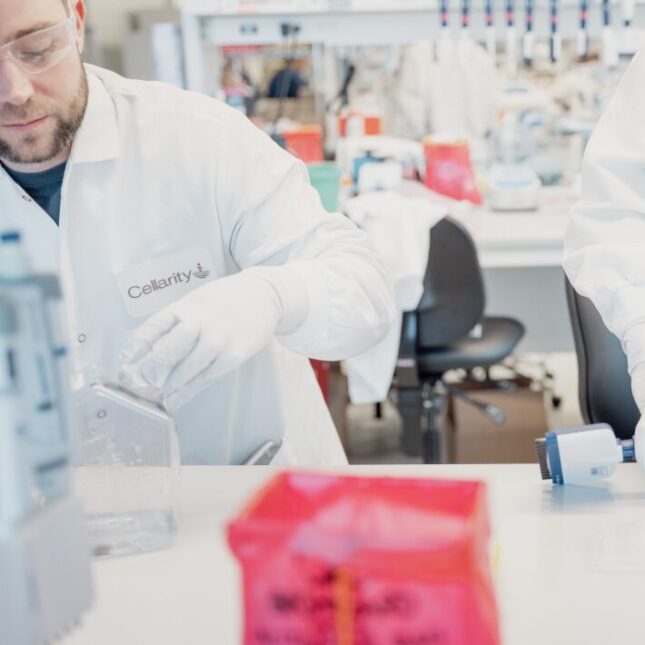 Two people in white lab coats using different equipment in a lab, with only the left person's face shown, wearing goggles. The logo of Cellarity, a pharmaceutical company, is displayed on their breast pocket— business coverage from STAT