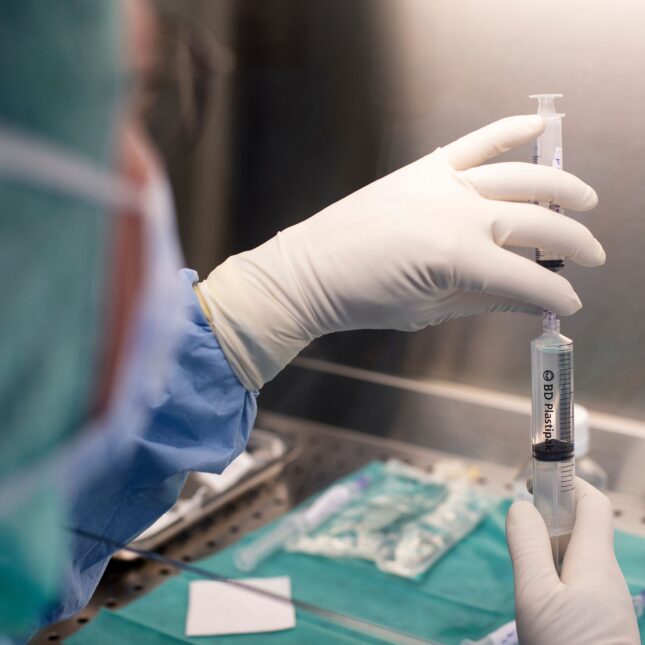 A pharmacist in a lab, sitting at a station, holds a larger syringe with their right hand and stacks a smaller syringe on top with their left hand, pulling a solution of phages from one to the other — first opinion coverage from STAT