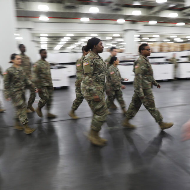 A group of ten military personnel in uniform marches forward through the grey floor of Javits Convention Center — health coverage from STAT