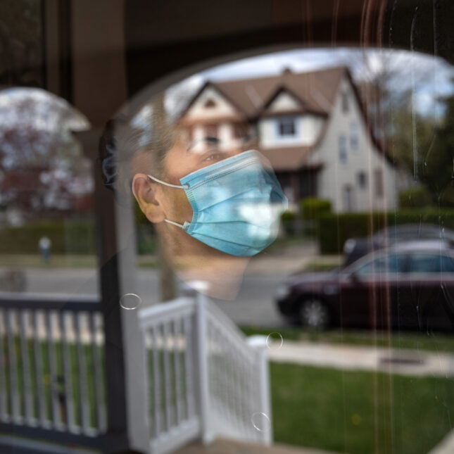 A masked person stands inside a house and behind a window that is reflecting the front yard and the houses across the street — health coverage from STAT