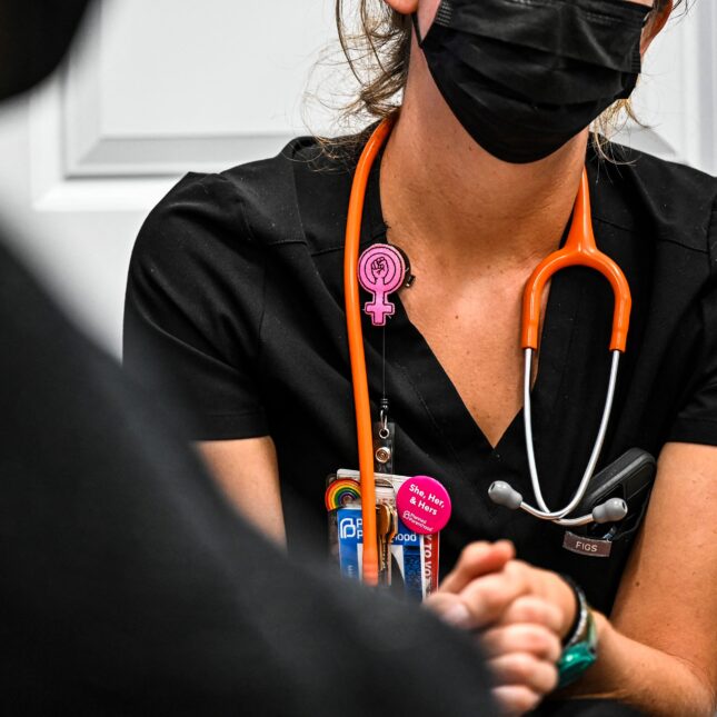 A nurse, wearing a rainbow pin, a pronoun badge, and a name badge holder featuring a fist icon inside a Venus symbol, sits across from a patient at Planned Parenthood Abortion Clinic in Jacksonville — health coverage from STAT