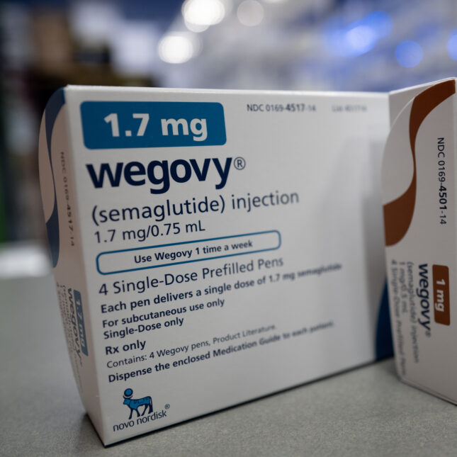 Two boxes of Wegovy stand together, with the 1.7 mg box in blue positioned slightly behind and to the left, and the 1 mg box in brown placed in front and to the right — first opinion coverage from STAT